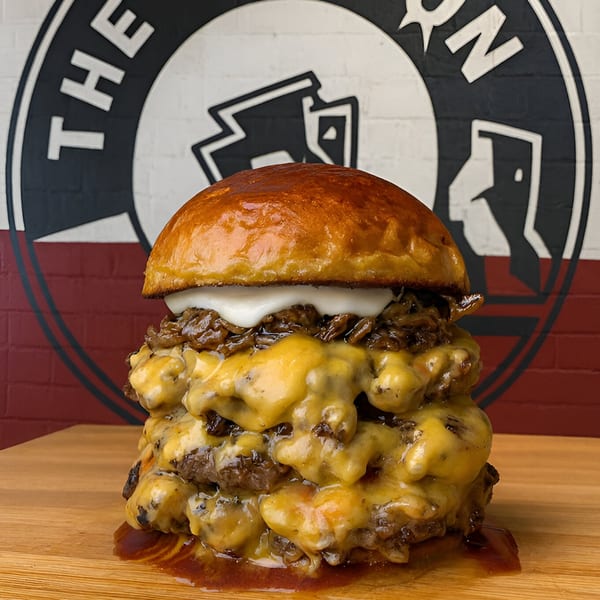 A towering triple cheeseburger from Refuel at The Station, with caramelized onions and white sauce, on a wooden board.